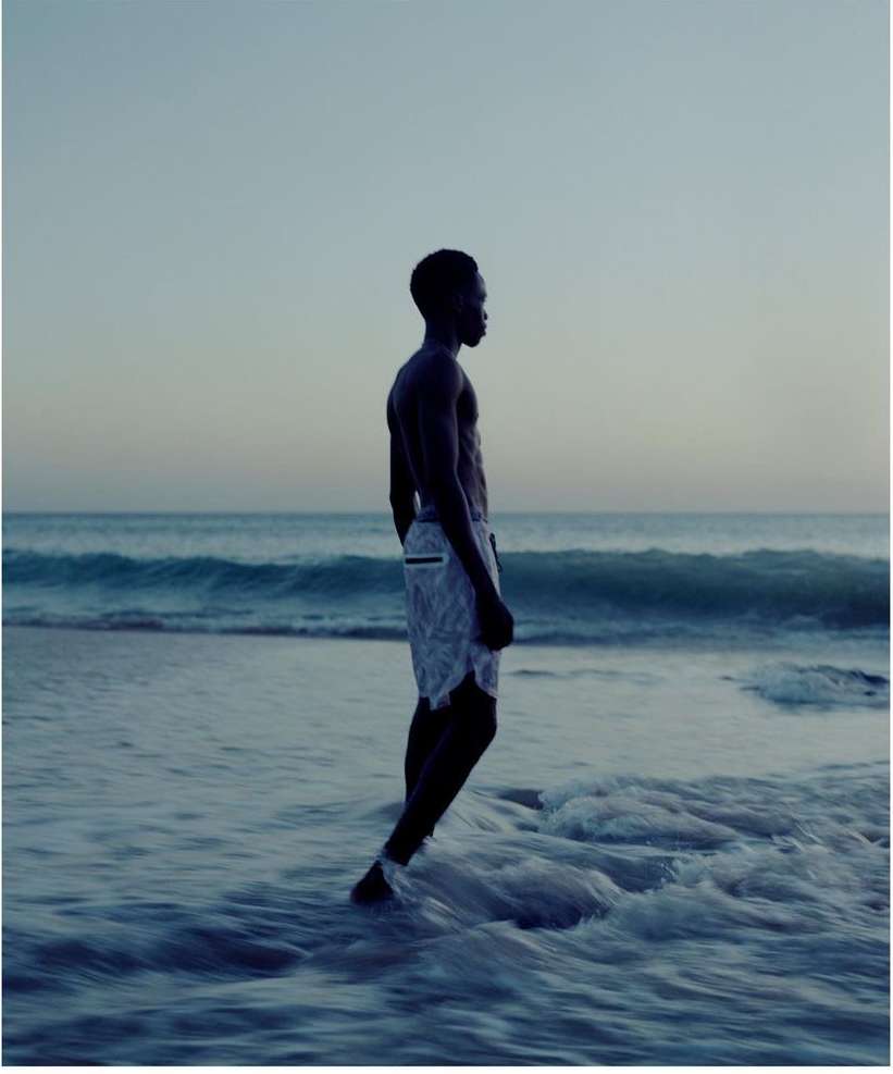 A slender, shirtless Black man stands in ocean water at the shoreline during daylight, side view, wearing shorts, against a sky backdrop.