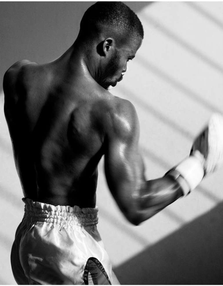 Monochrome portrait of a muscular Black man in boxing shorts, back to camera, wearing boxing gloves, posed in soft, even lighting.
