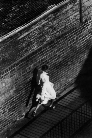 High-angle monochrome photograph of a person walking beside a brick wall in London, casting a long shadow.