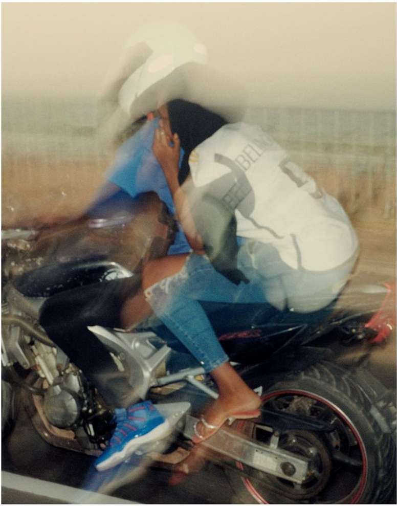Two people ride a motorcycle on a blurred road, the passenger wearing a helmet, blue jeans, and gold sandals, with motion blur.
