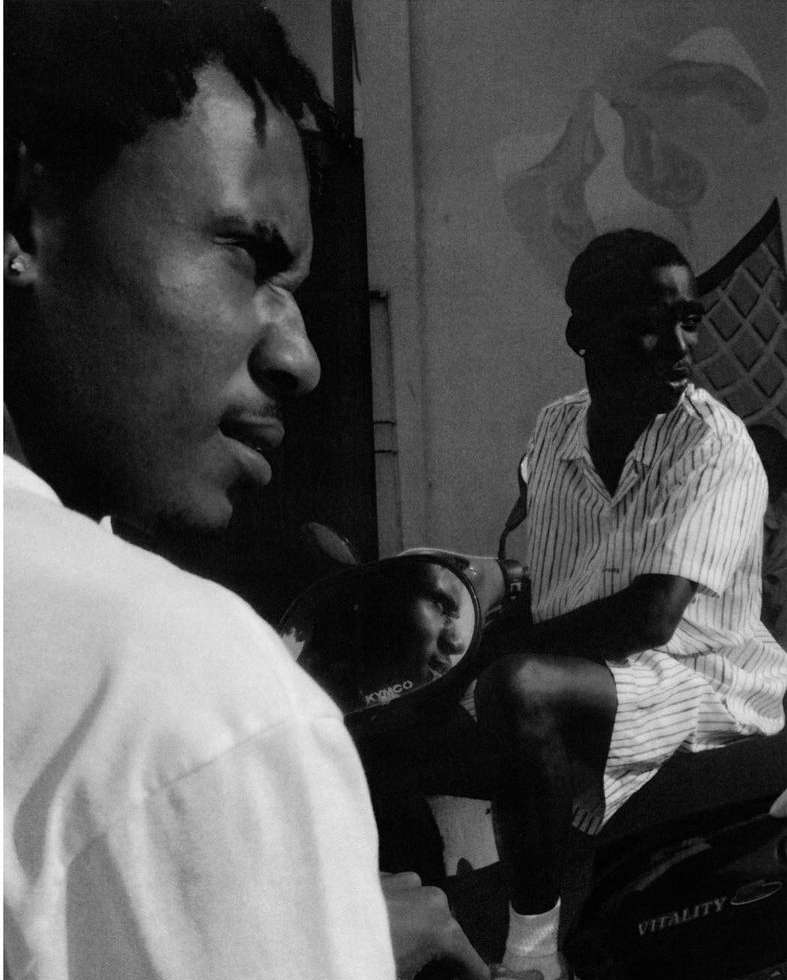 Black and white medium shot of two dark-skinned men outdoors, one with dreadlocks on a scooter with visible KYMCO logo.