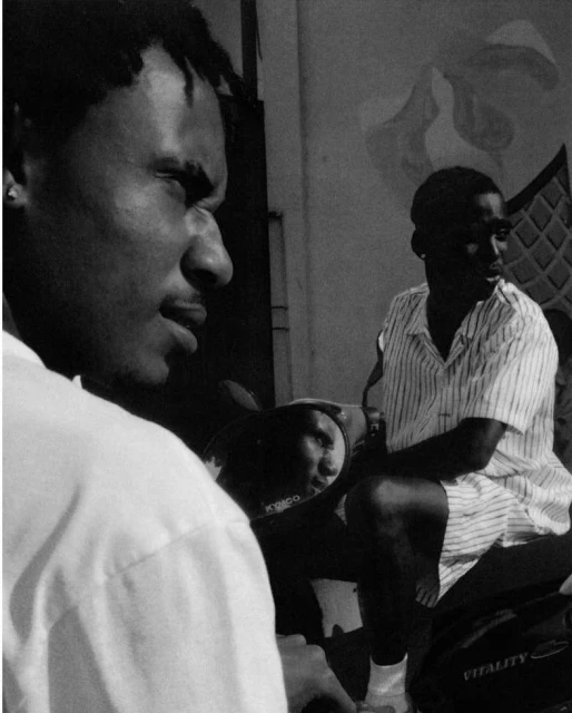 Black and white medium shot of two dark-skinned men outdoors, one with dreadlocks on a scooter with visible KYMCO logo.