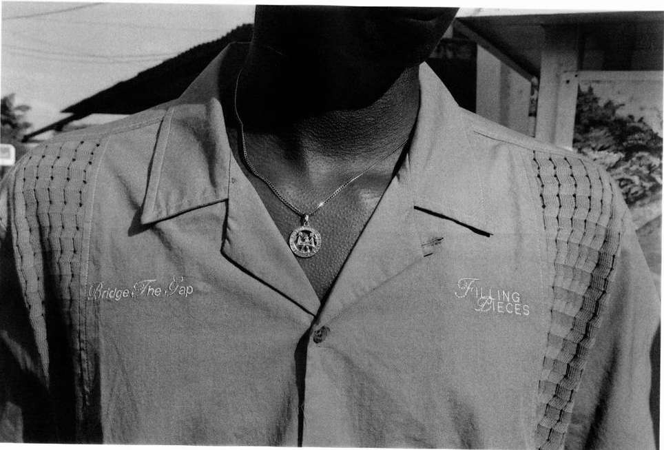 Close-up of a person wearing a button-down shirt with embroidered text and a pendant necklace in a black and white photo.
