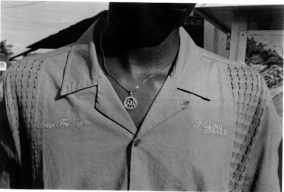 Close-up of a person wearing a button-down shirt with embroidered text and a pendant necklace in a black and white photo.