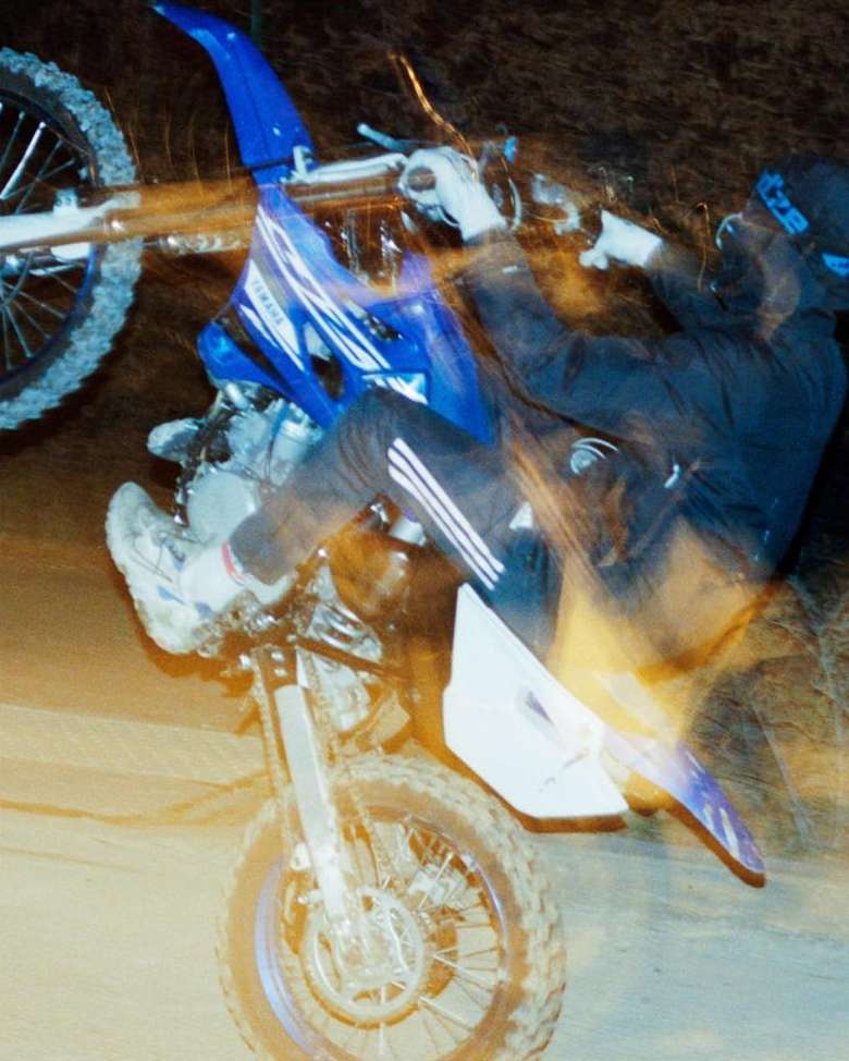 Motocross rider performs a wheelie at night; blue dirt bike with white accents; athletic male wearing black and white riding gear.