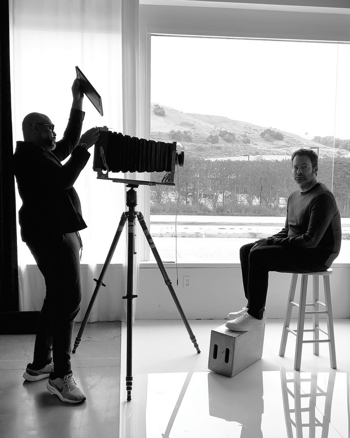Black and white studio shot of photographer using a large format camera to photograph a seated adult male, with a bright outdoor background.