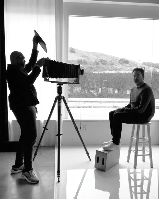 Black and white studio shot of photographer using a large format camera to photograph a seated adult male, with a bright outdoor background.