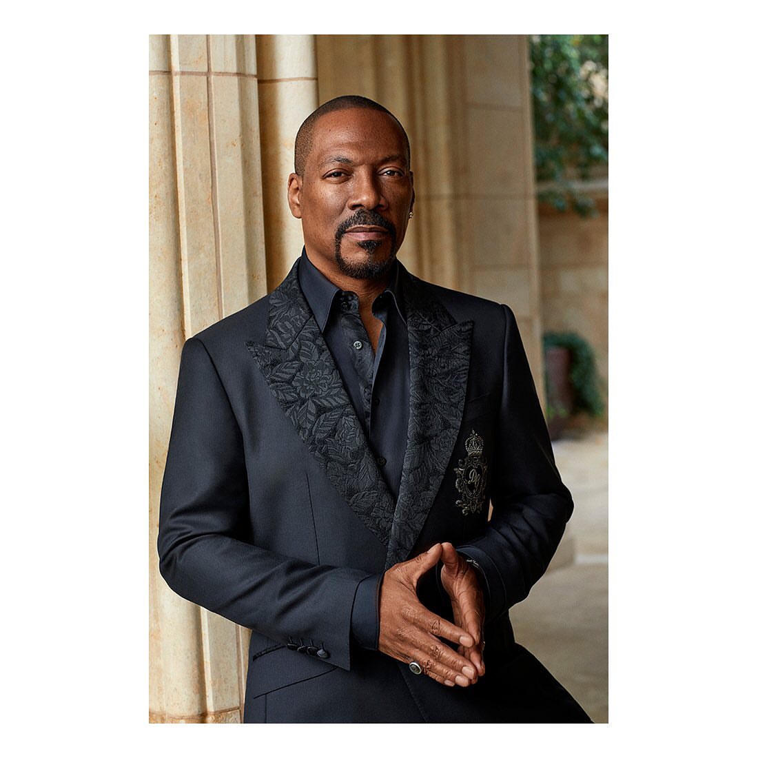 A dignified portrait of an African-American man in a black suit with embroidered details, standing against a neutral-toned architectural backdrop.