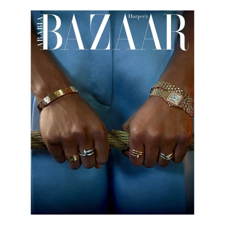 Close-up of hands wearing gold bangles, rings, and a luxury watch while gripping a thick textured rope against a blue suit.