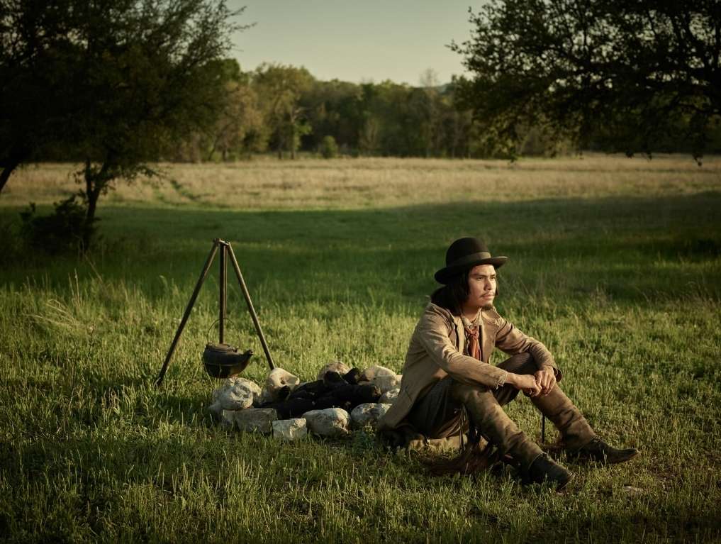 Person in traditional clothing sits near campfire in grassy field with trees. Pot hangs on tripod over the fire under natural lighting.