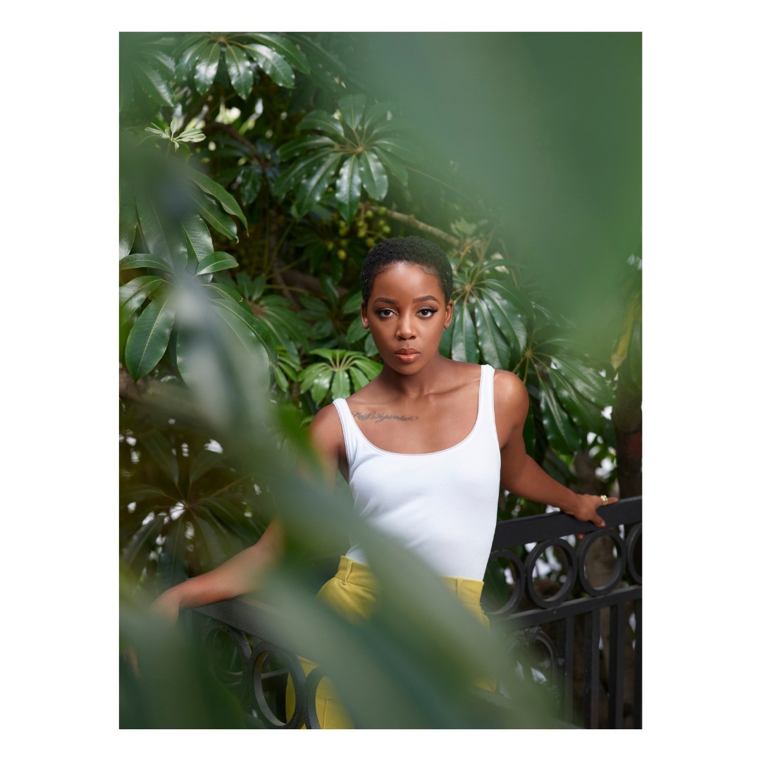 Portrait of a young Black woman with short hair posing outdoors, framed by green foliage, wearing a white tank top and yellow pants.