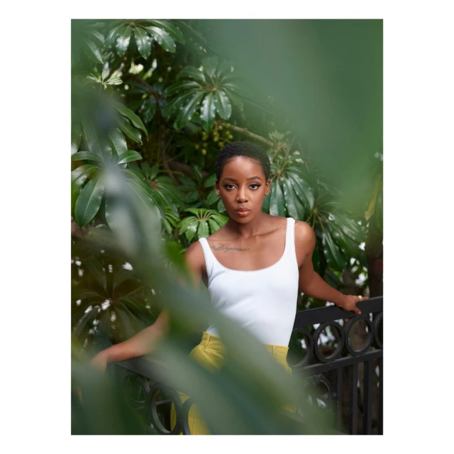 Portrait of a young Black woman with short hair posing outdoors, framed by green foliage, wearing a white tank top and yellow pants.