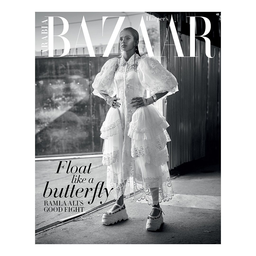 Black and white magazine cover featuring a woman in a white dress and platform shoes, standing against an industrial backdrop.