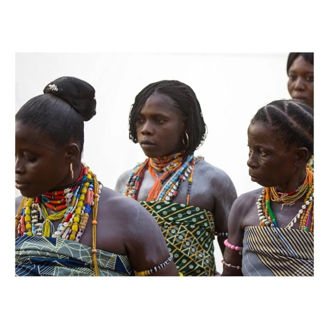 Three dark-skinned women adorned with colorful beaded necklaces and patterned fabrics, stand together in an outdoor setting with a bright background.