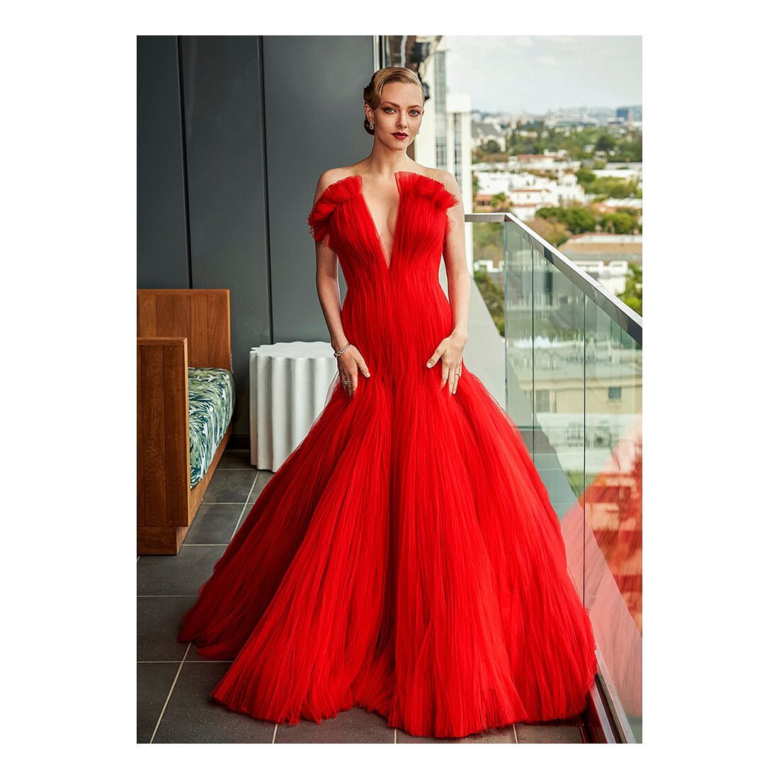 Amanda Seyfried poses on a balcony wearing a red, floor-length gown. The urban skyline is seen in the background during the daytime.
