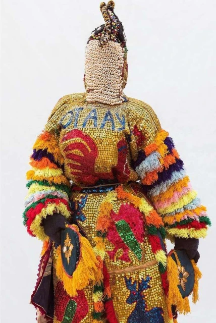 Full-length studio shot of an individual wearing a colorful, ornate costume with animal motifs and beaded details against a white backdrop.