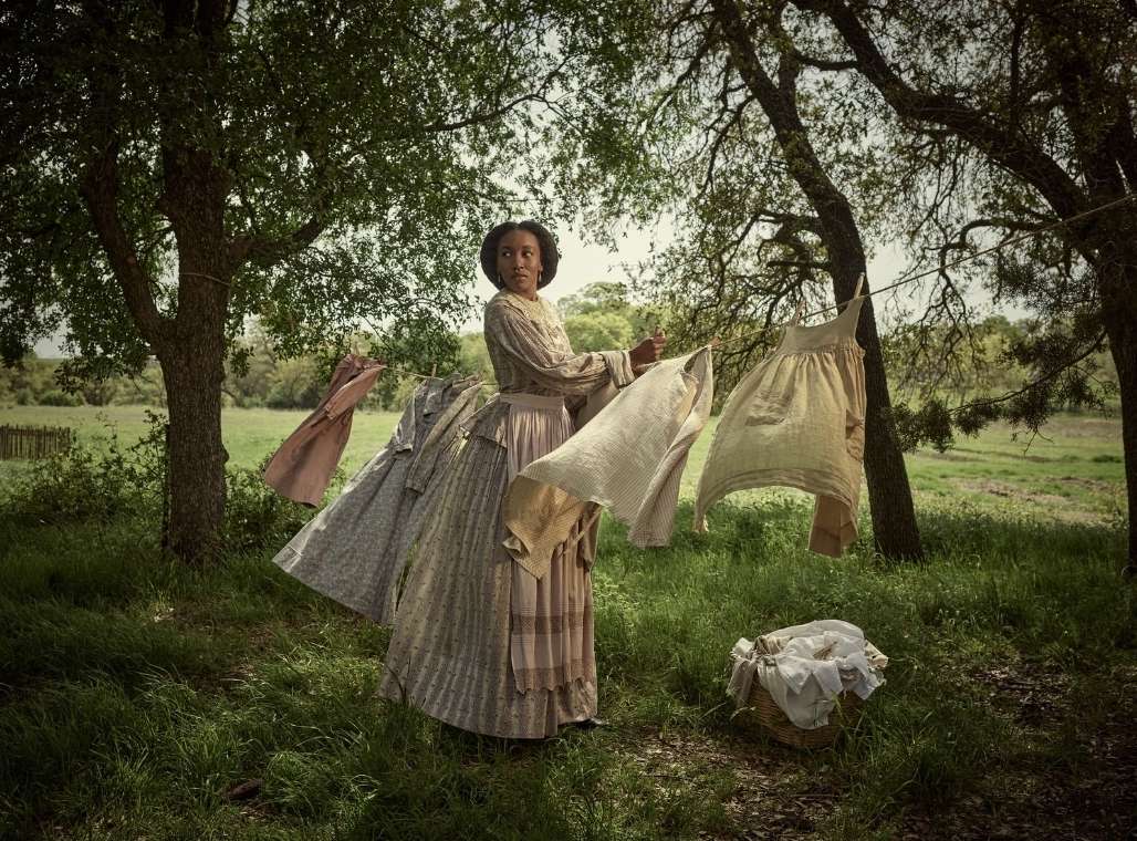 An African-American woman in period clothing hangs laundry to dry on a clothesline strung between trees in a grassy, rural setting.