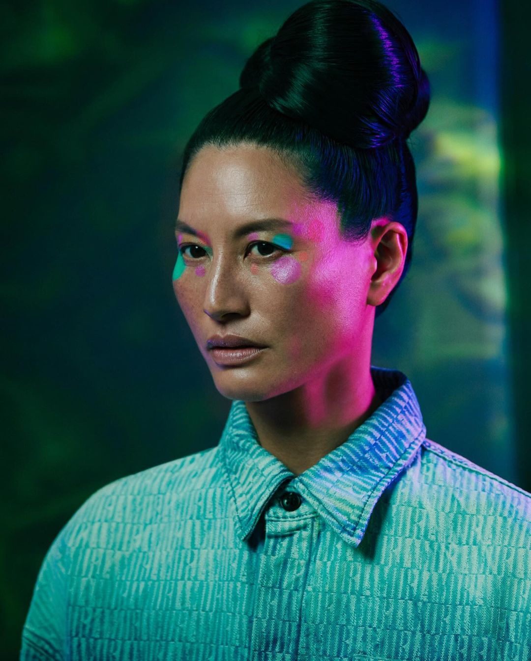 Woman with creative teal and glitter makeup poses in front of a green and blue background. She wears a textured blue shirt.
