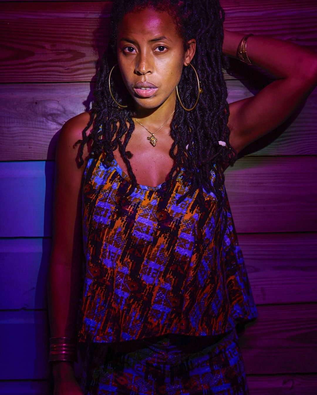 Woman with dreadlocks wears a patterned top and gold jewelry. She is posing in front of a wooden wall with purple and red lighting.
