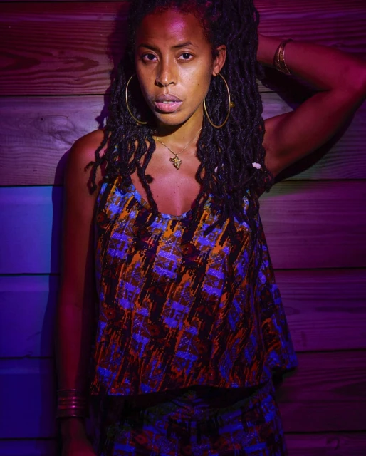 Woman with dreadlocks wears a patterned top and gold jewelry. She is posing in front of a wooden wall with purple and red lighting.