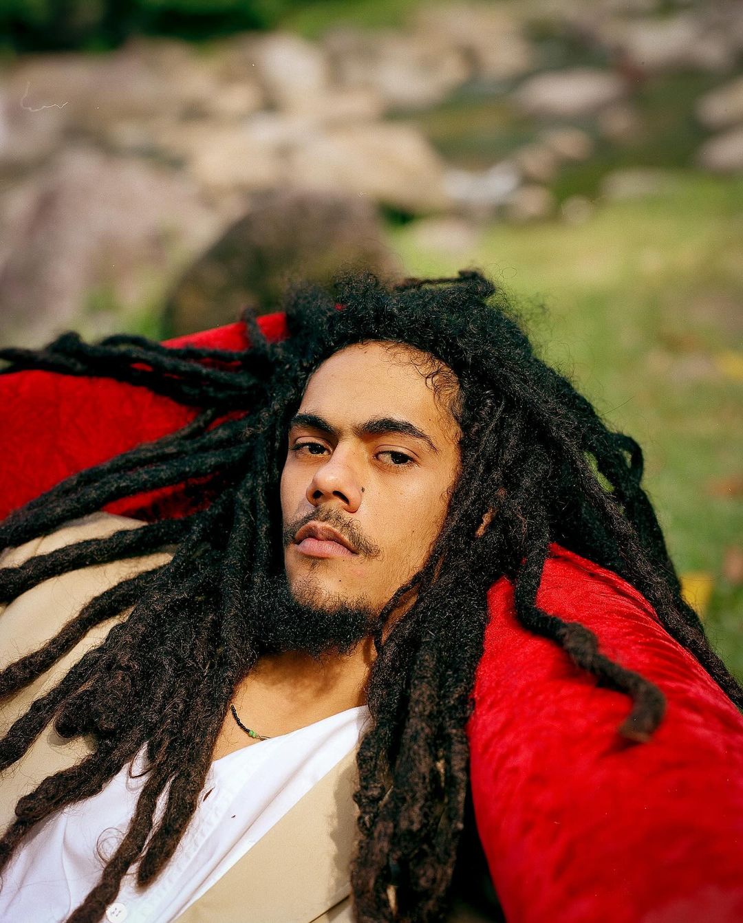 Portrait of a man with long dreadlocks and a slight mustache wearing a white shirt and tan jacket in an outdoor setting.