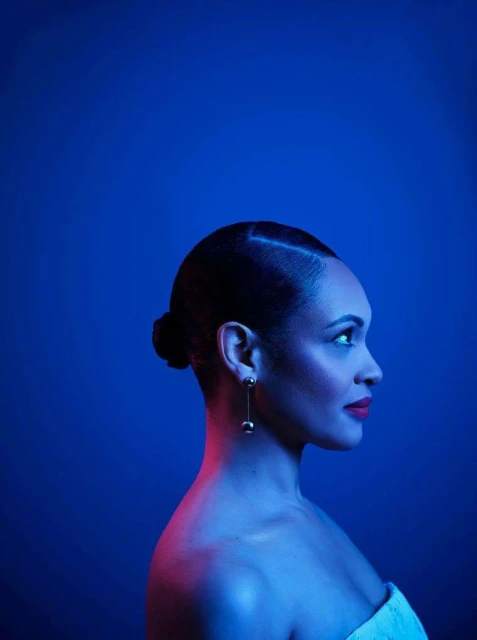 Profile view of a stylish woman with a sleek updo wearing dangling ball earrings against a solid blue background with red lighting.