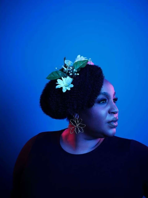 Portrait of an adult woman with an afro hairstyle decorated with white flowers, wearing flower earrings and a black top in a blue environment.