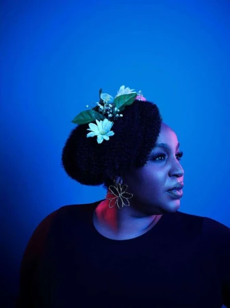 Portrait of an adult woman with an afro hairstyle decorated with white flowers, wearing flower earrings and a black top in a blue environment.