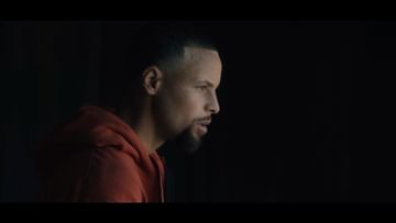 Close-up shot of Stephen Curry in a dimly lit setting, wearing a red hooded sweatshirt, captured with shallow depth of field. Focus on his profile.