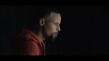 Close-up shot of Stephen Curry in a dimly lit setting, wearing a red hooded sweatshirt, captured with shallow depth of field. Focus on his profile.