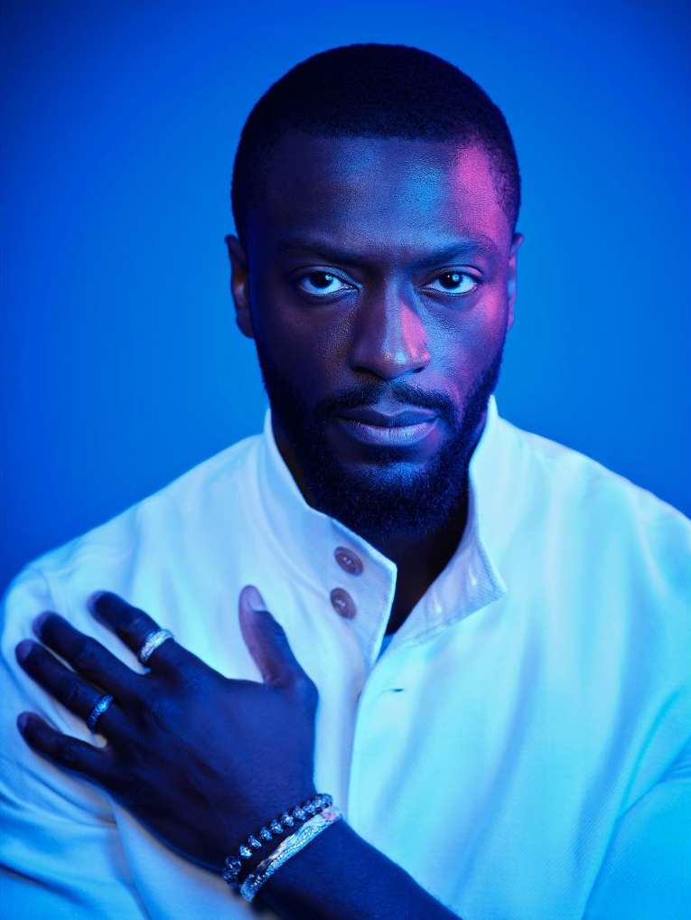 Portrait of a Black man with a beard wearing a white buttoned shirt against a blue background