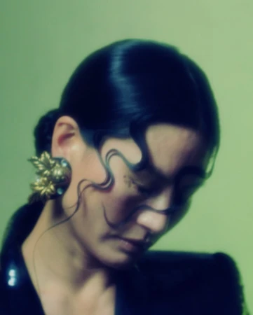 Portrait of a woman with finger waves framing her face, wearing ornate gold leaf earrings against a muted green background.