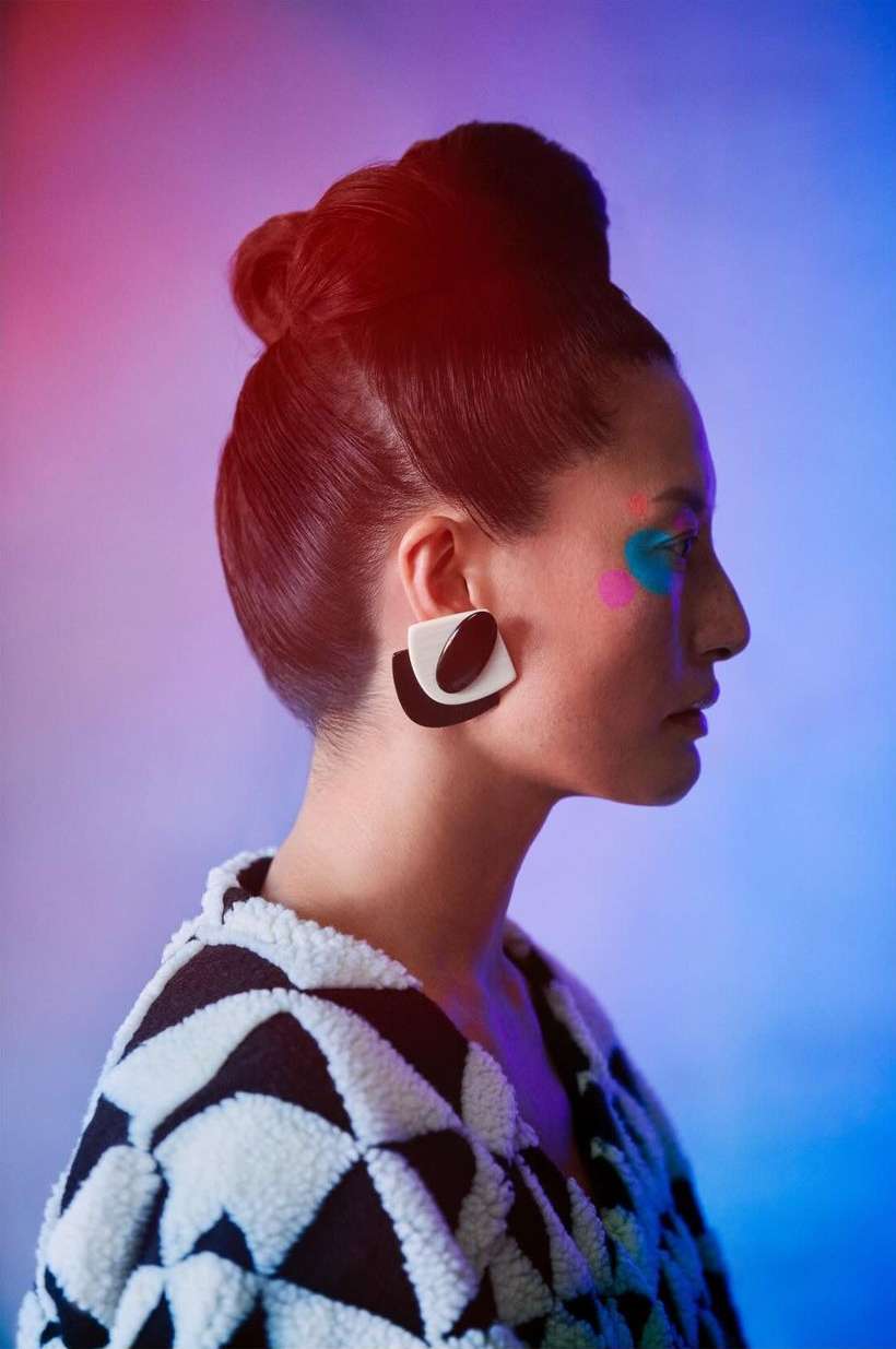 Profile shot of a woman with high bun hairstyle, makeup accents, black and white earrings, and black and white patterned coat.