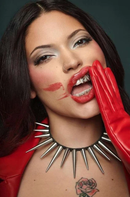 Brunette adult female with lipstick smudge on her face wears a spiked collar, red jacket, and gloves while holding her hand to her mouth.