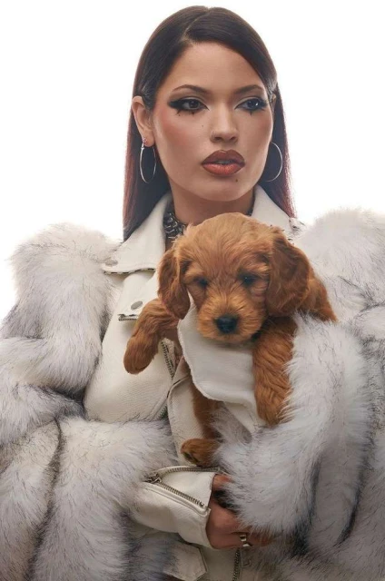 Portrait of a woman with dramatic makeup holding a brown puppy wearing a fur coat in a bright studio environment.