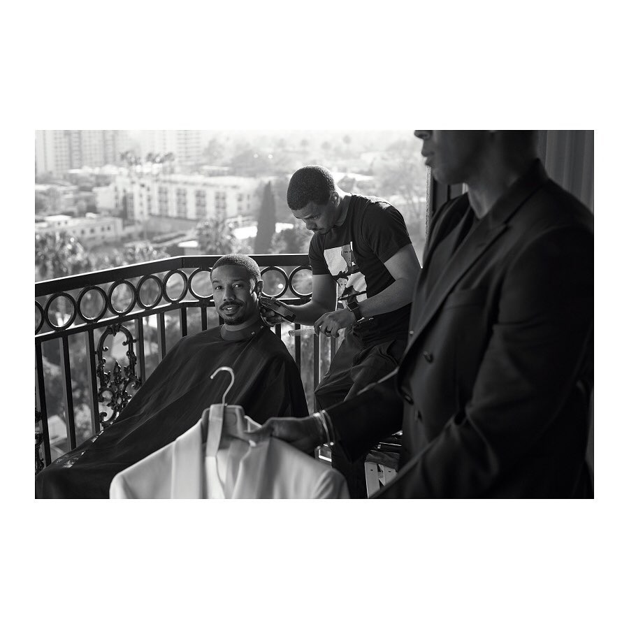 Michael B. Jordan gets a haircut on a balcony overlooking the city, assisted by a barber and a man holding a jacket.