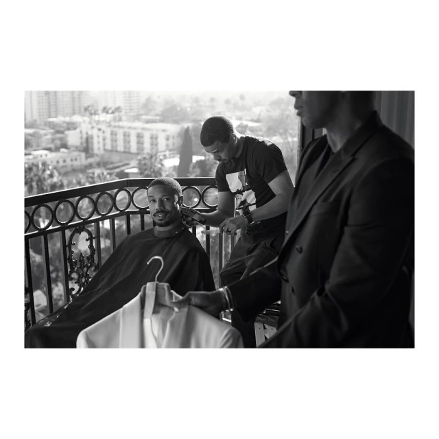 Michael B. Jordan gets a haircut on a balcony overlooking the city, assisted by a barber and a man holding a jacket.