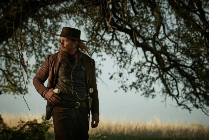 Cowboy in a brown coat and hat standing outdoors under tree branches with a grassy field background