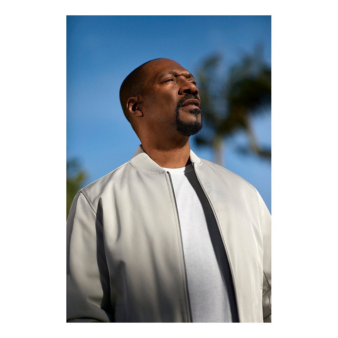 Outdoor portrait of a man with a beard wearing a white jacket, looking up towards the sky with trees and blue sky in the background.
