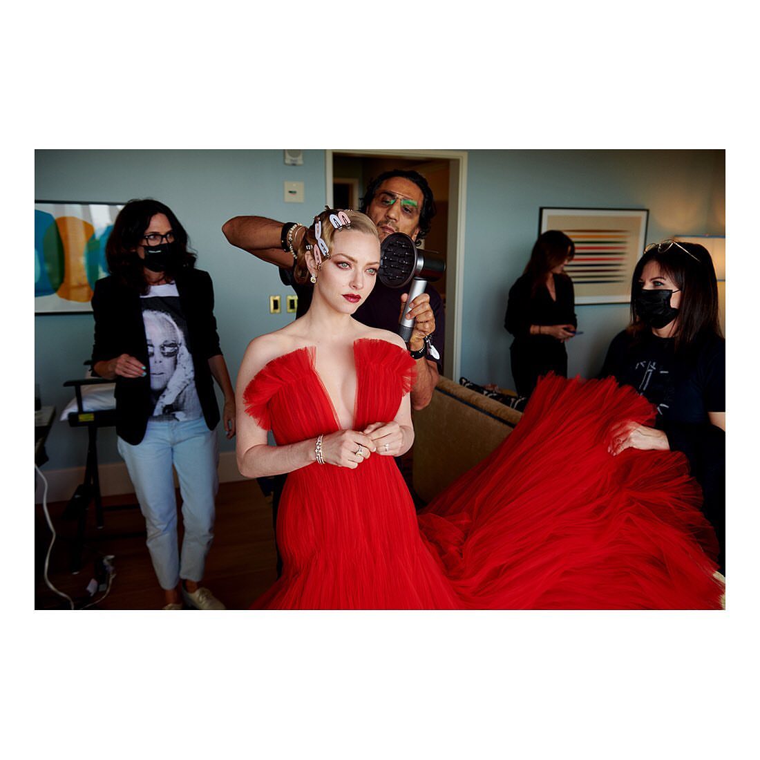 Amanda Seyfried in red tulle gown getting hair styled indoors, photo studio setting, stylists, dress with train, celebrity event prep.