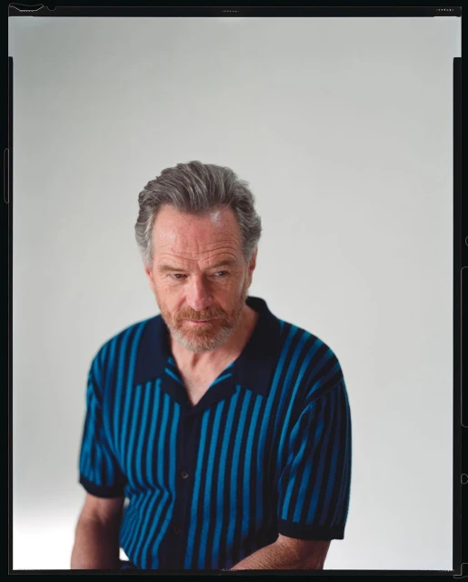 Portrait of a mature adult male with a short beard wearing a blue and black striped polo shirt, looking slightly downward against a plain white backdrop.