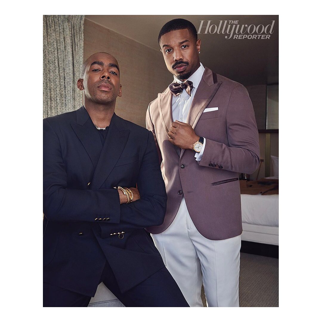 Two adult men pose in a photo studio, one in a dark suit with his arms crossed, the other in a mauve suit with The Hollywood Reporter logo.