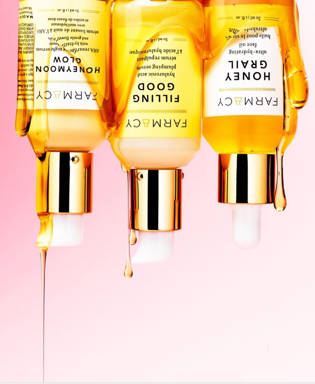 Three FARMACY serum bottles stand against a pink background, with drops of golden serum dripping from the bottles' gold and white pumps.