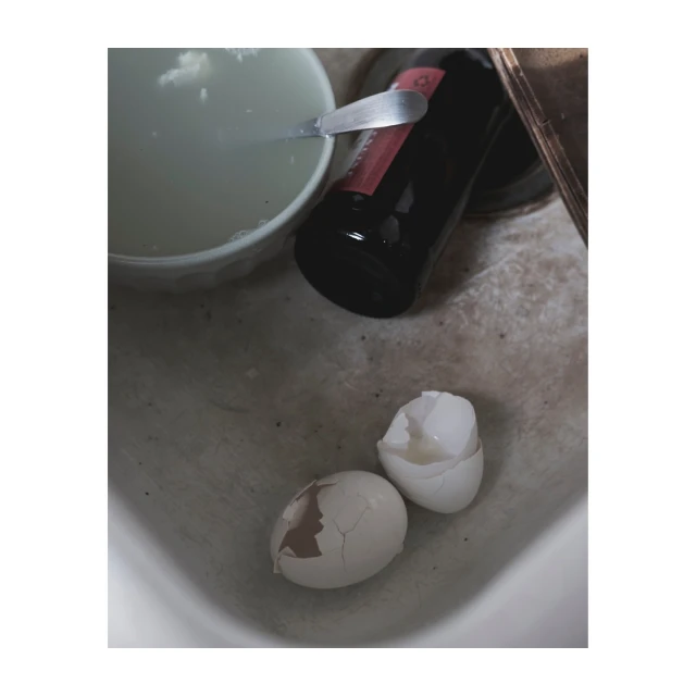 Overhead shot of two cracked eggs, a bowl, and a bottle on a concrete surface, lit with muted, natural light, creating a simple still life.