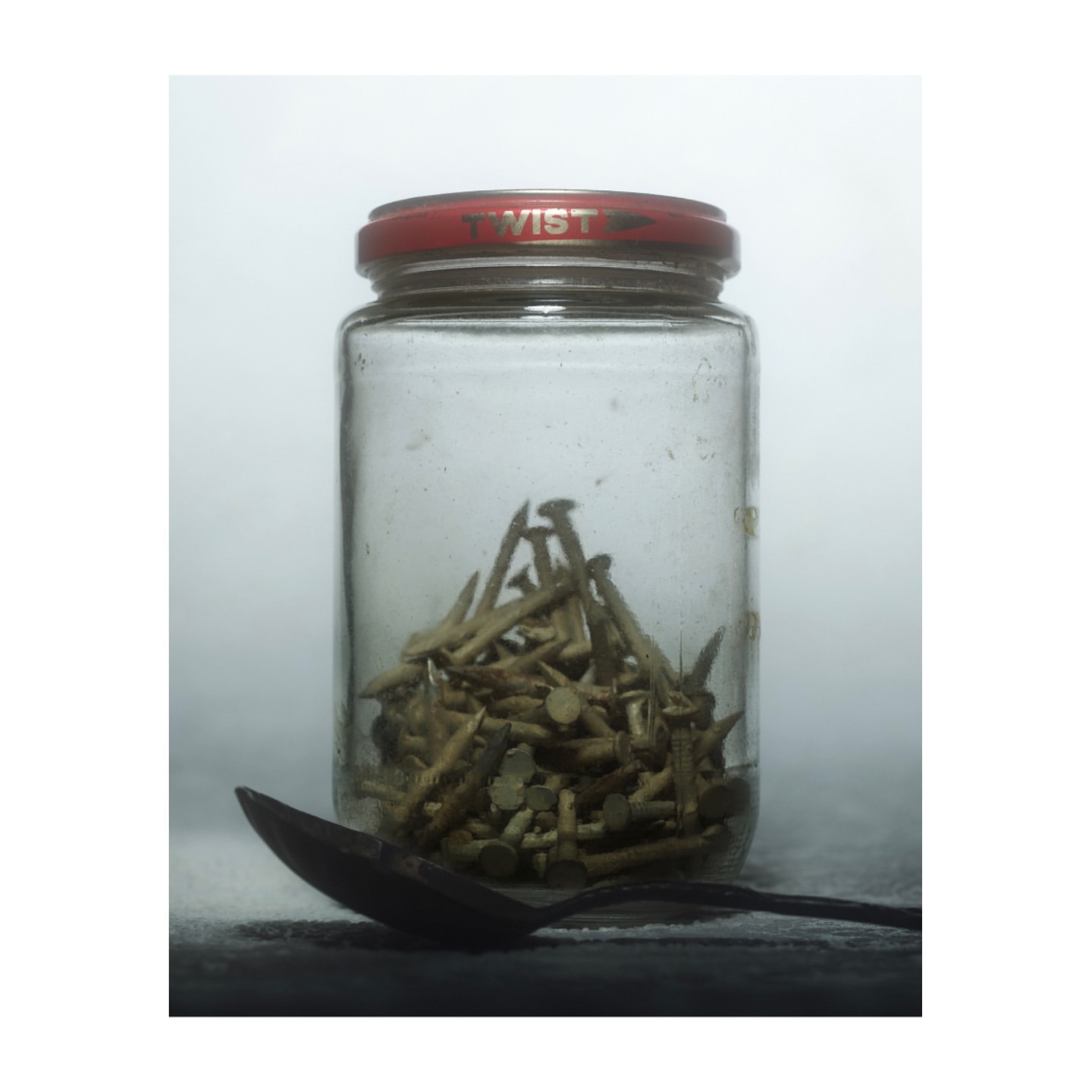 Still life photograph of a glass jar filled with old nails, next to a vintage metal spoon, on a dark surface against a white background.