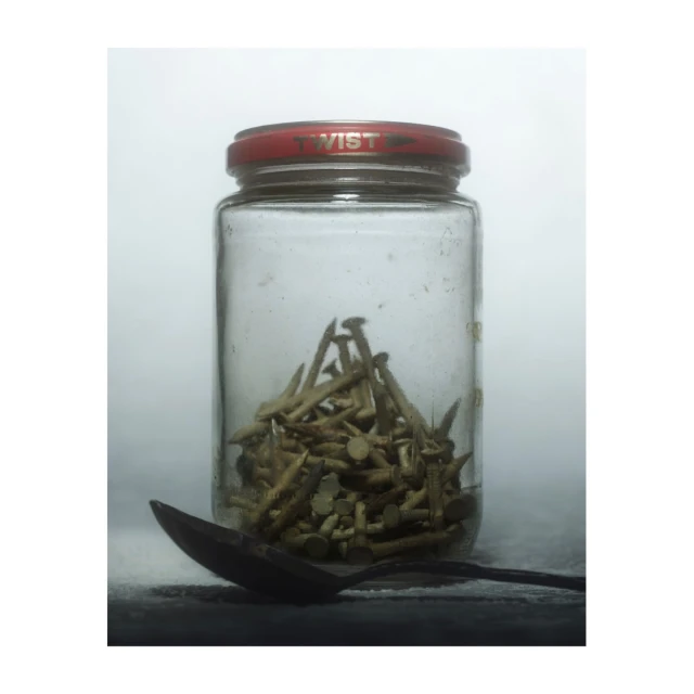 Still life photograph of a glass jar filled with old nails, next to a vintage metal spoon, on a dark surface against a white background.