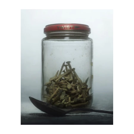 Still life photograph of a glass jar filled with old nails, next to a vintage metal spoon, on a dark surface against a white background.