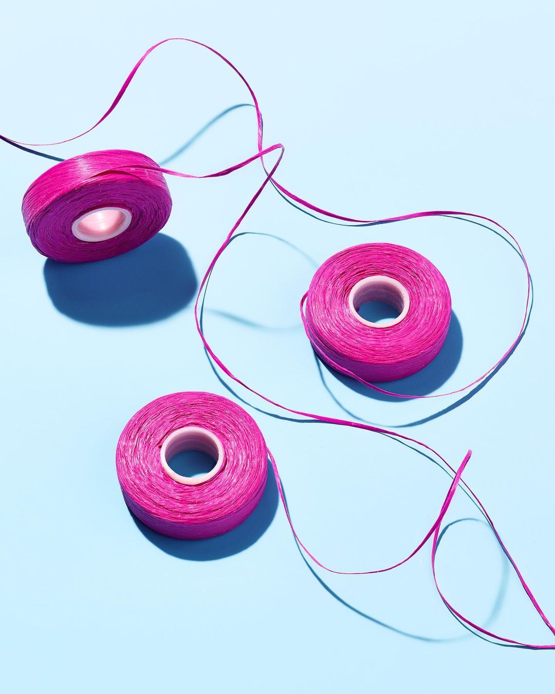 Three spools of vibrant pink thread are arranged on a soft blue background. The composition features the flowing thread and shadows.