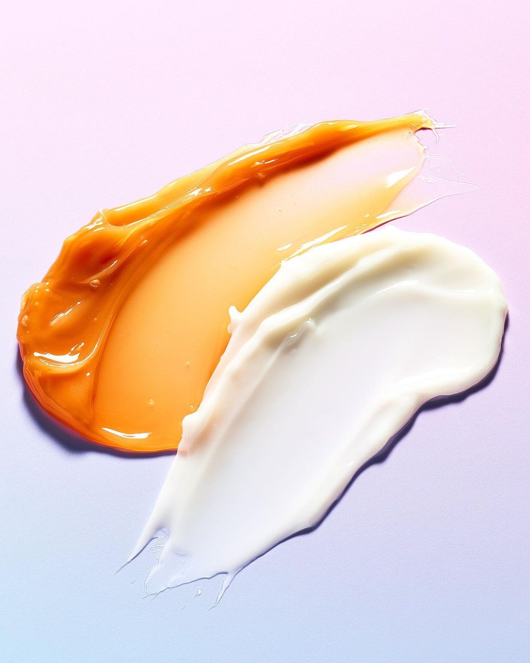 Close-up of smears of amber and white cosmetic creams against a soft gradient pink and blue background. Beauty product shot.