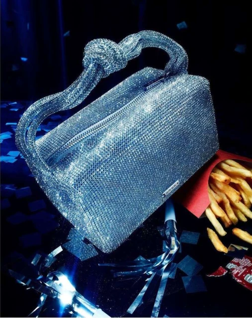 Sparkly silver handbag with knot handle, set with French fries and silver streamers on a dark background. Fashionable accessorization.